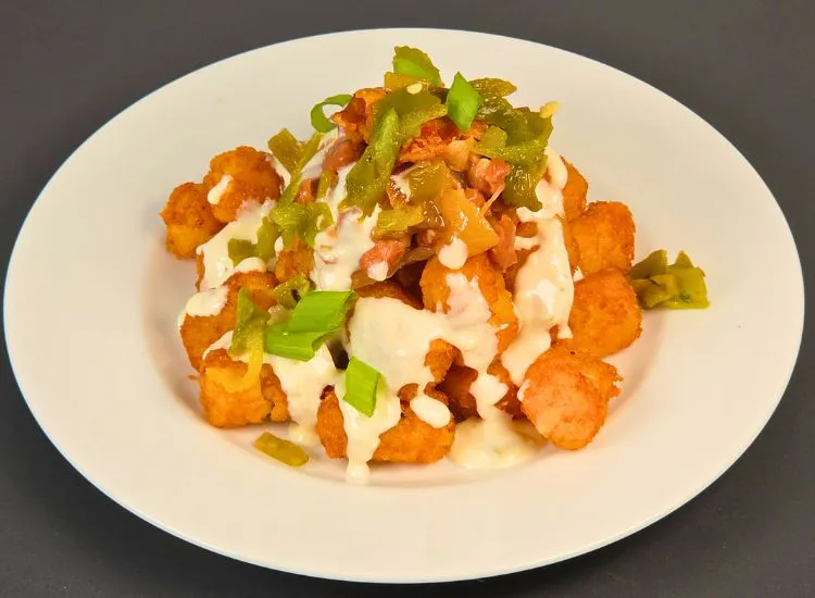 Crispy tater tots covered in creamy sauce, chopped hatch chiles, bacon, and green onions on a white plate.
